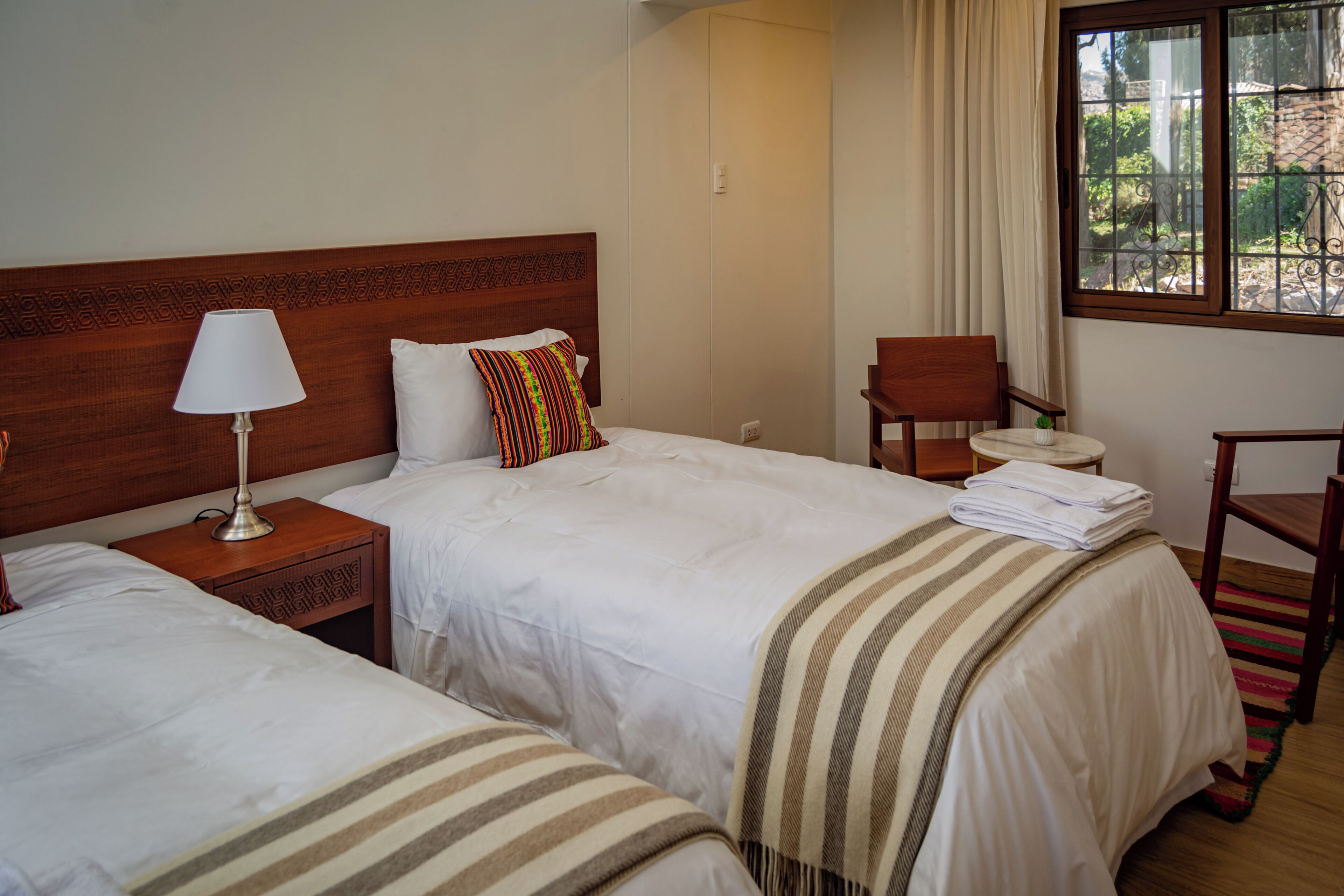 A hotel room with two beds, a wooden headboard, a nightstand with a lamp, a chair, and a window with a view of greenery.