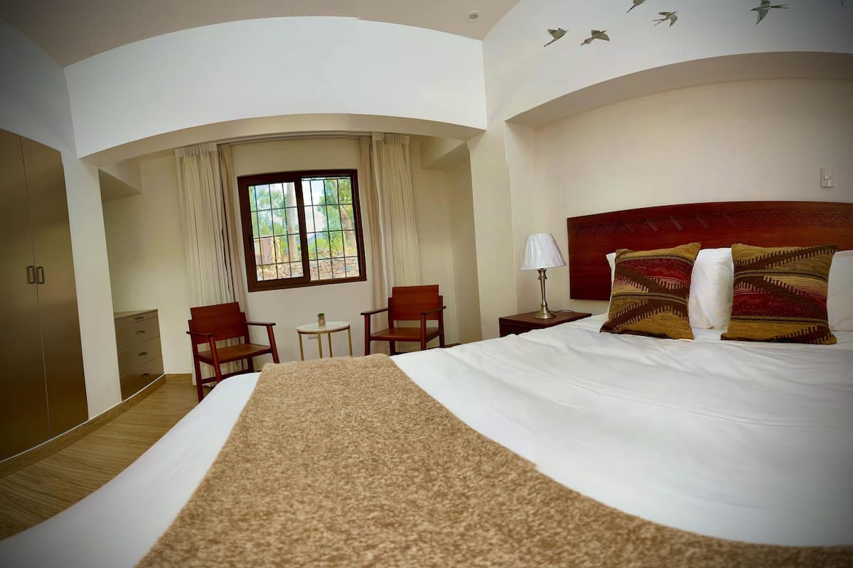 Habitación de hotel con una cama grande, cabecera de madera, escritorio, silla, mesita y una ventana con cortinas.