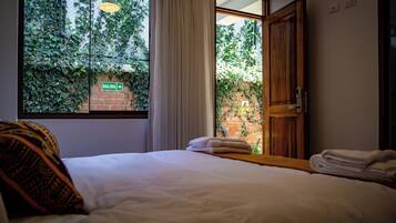 Habitación de hotel con cama, ventana con cortinas, puerta y vista a un muro de ladrillo con vegetación.