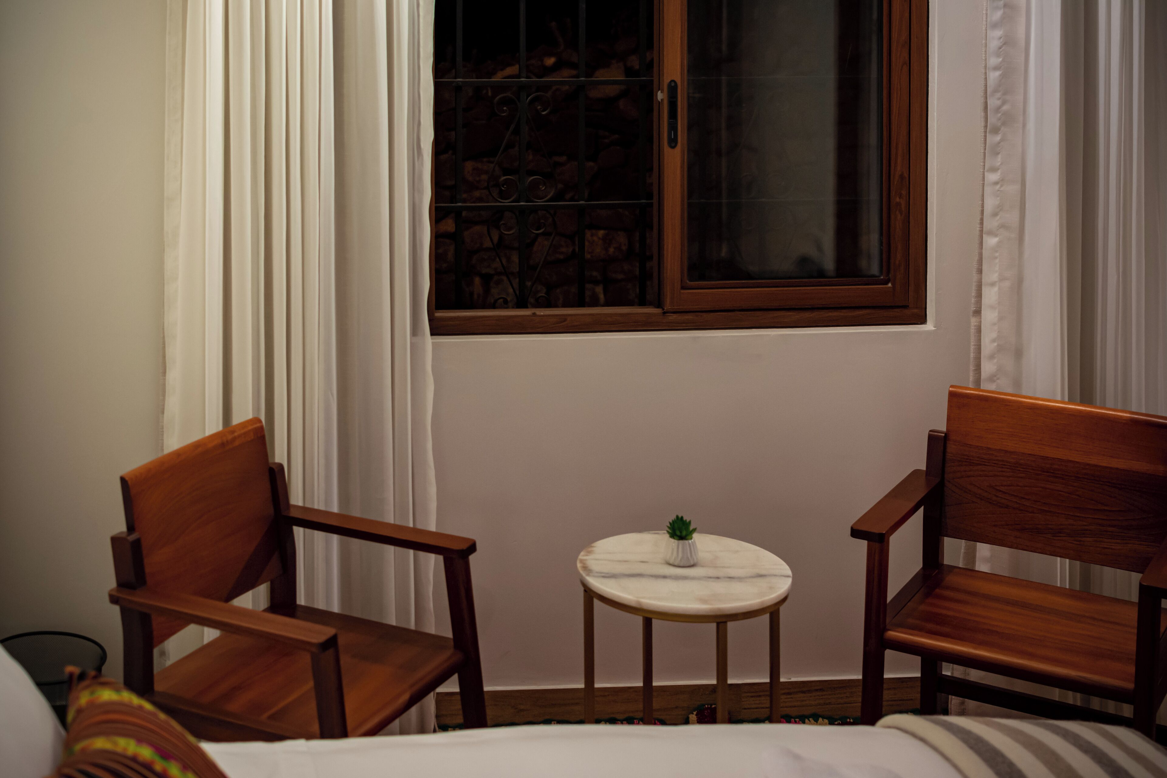A room with two wooden chairs, a small round table with a plant, and a window with a view of a stone wall.