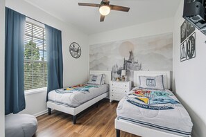 Un dormitorio con dos camas, un ventilador de techo, una ventana con cortinas, un cuadro colgado en la pared y una mesita de noche con una lámpara.