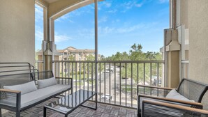 A balcony with a sofa and chairs, a view of a parking lot, and buildings in the background.