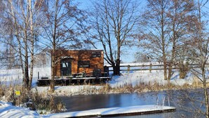 A cabin with a deck overlooking a frozen pond, surrounded by trees and a wooden fence. - Tiny House by a swimming pond with terrace & fireplace – Last Minute (Großharthau)