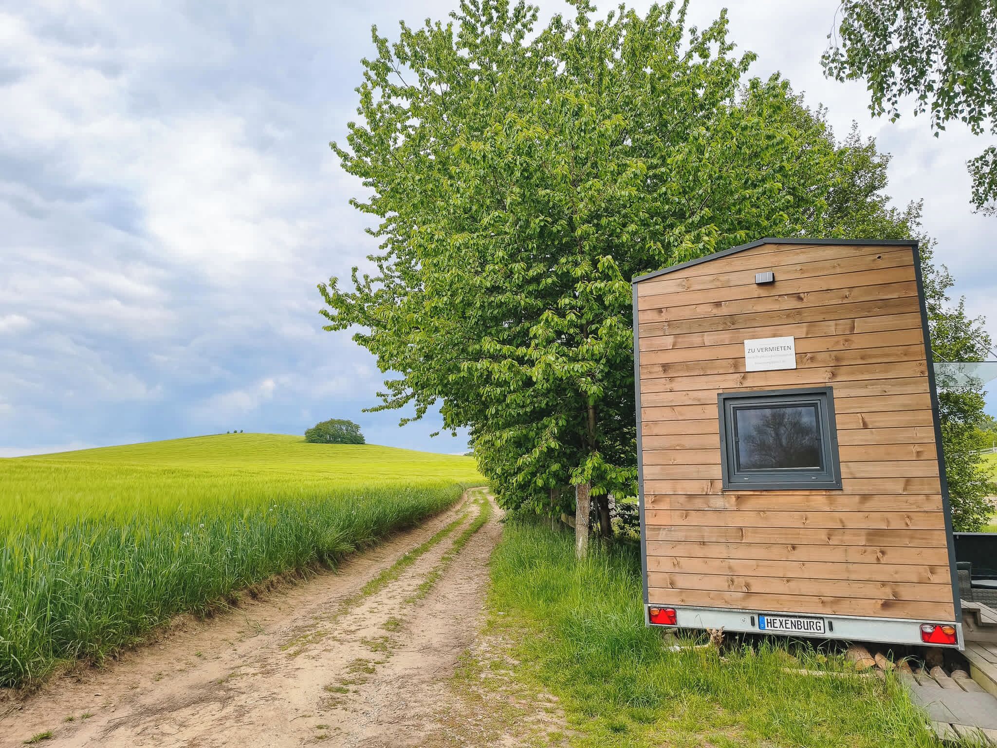 Ein Holz-Wohnmobil mit Fenster und einem Nummernschild mit der Aufschrift 'HEXENBURG' steht neben einer unbefestigten Straße, mit einem grünen Feld und einem großen Baum im Hintergrund.