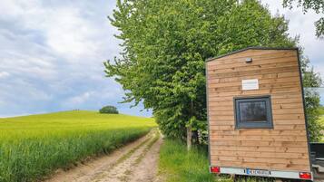Ein Holz-Wohnmobil mit Fenster und einem Nummernschild mit der Aufschrift 'HEXENBURG' steht neben einer unbefestigten Straße, mit einem grünen Feld und einem großen Baum im Hintergrund.