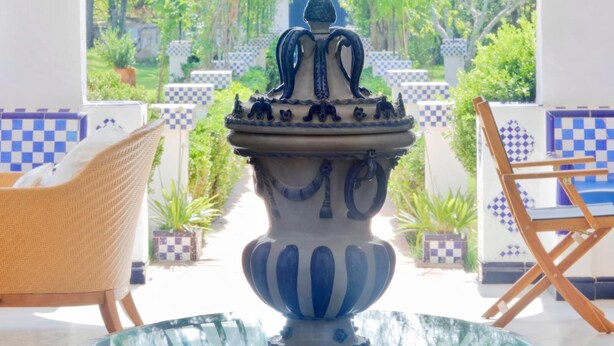 Eine dekorative, keramische Vase mit blau-weißem Muster, platziert auf einem Glastisch in einem sonnendurchfluteten Raum mit Schachbrettfliesen und Korbmöbeln.