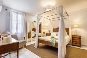 Una cama con dosel sobre cuatro postes, un cabecero de madera, una mesita de noche con lámpara, una silla y un espejo.