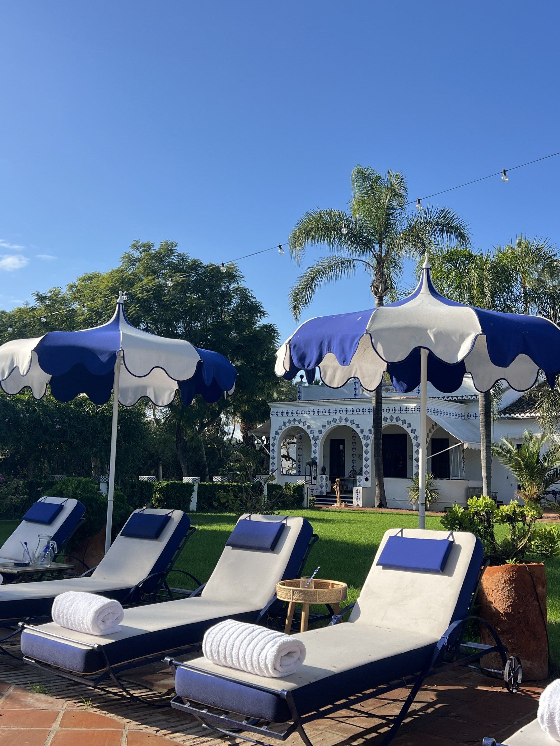 Un espace aménagé au bord de la piscine, avec des chaises longues et des parasols, un bâtiment orné d’arches et des palmiers en arrière-plan.