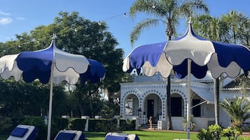Un espace aménagé au bord de la piscine, avec des chaises longues et des parasols, un bâtiment orné d’arches et des palmiers en arrière-plan.
