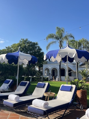 A poolside area with lounge chairs and umbrellas, a building with arches, and palm trees in the background. - 16th-century villa for 14, near Valencia | Villa Paula (Manises)
