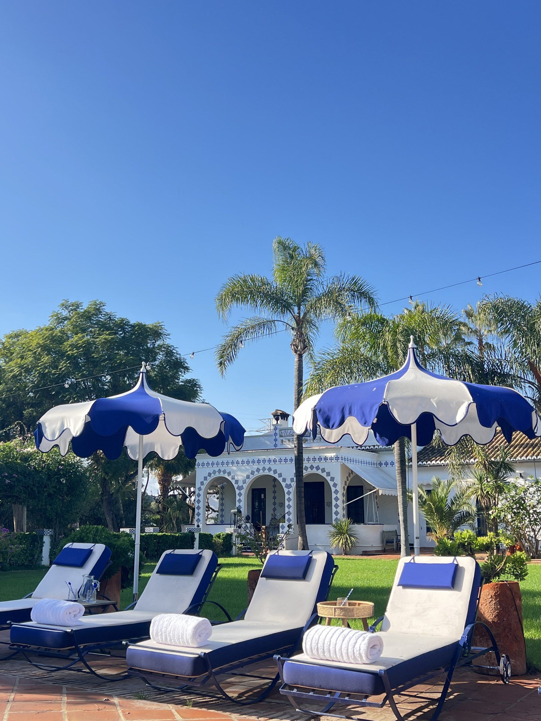 Zona de piscina con sillones de descanso y sombrillas, un edificio con arcos y palmeras al fondo.