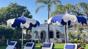 Un espace piscine avec des chaises longues et des parasols, un bâtiment orné d’arches et, au fond, des palmiers.