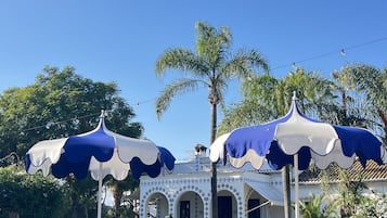 Zona de piscina con sillones de descanso y sombrillas, un edificio con arcos y palmeras al fondo.