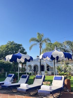 Zona de piscina con sillones de descanso y sombrillas, un edificio con arcos y palmeras al fondo.