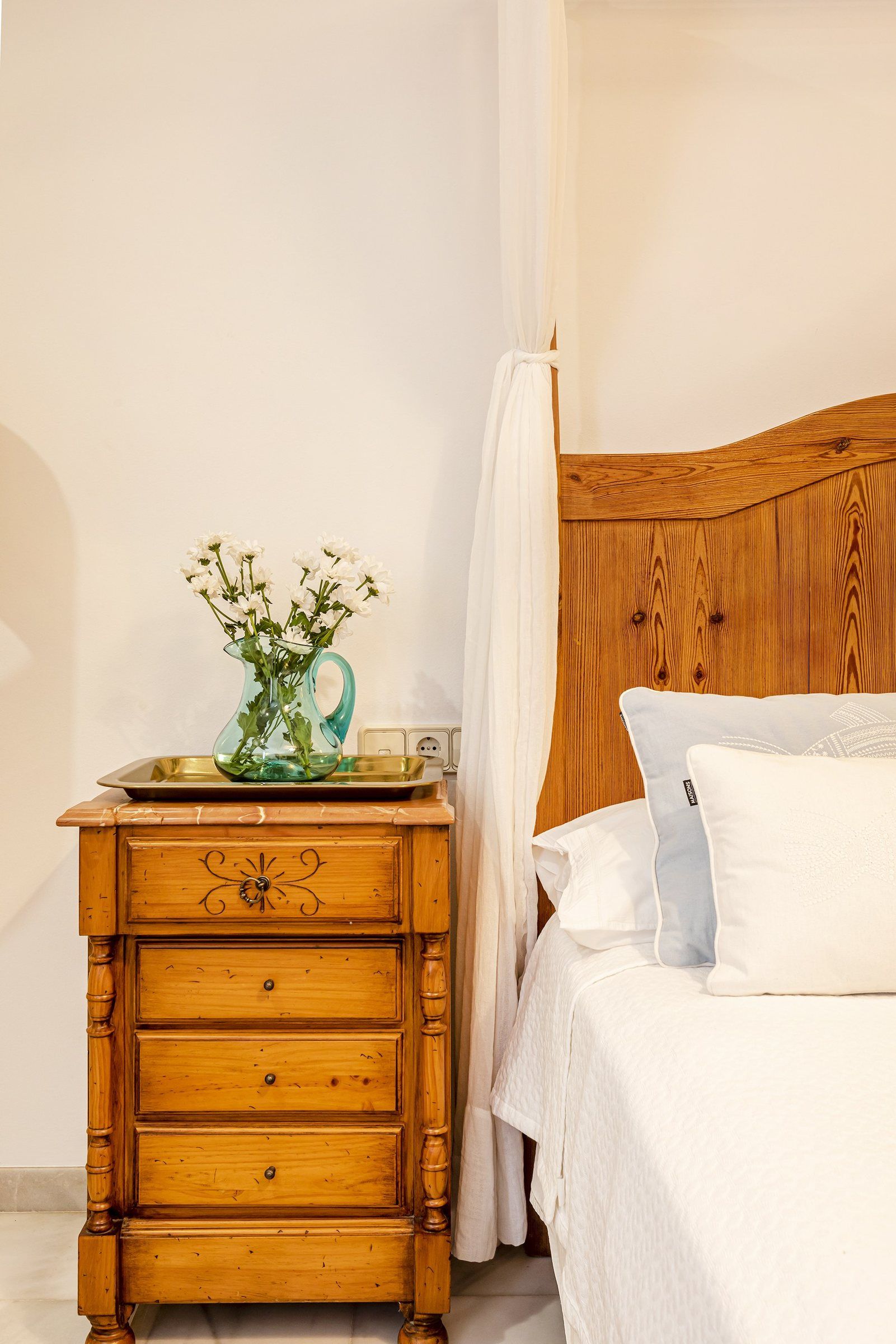Une table de chevet en bois avec des tiroirs, un vase en verre avec des fleurs, une table de chevet, un lit avec une literie blanche et une tête de lit en bois.