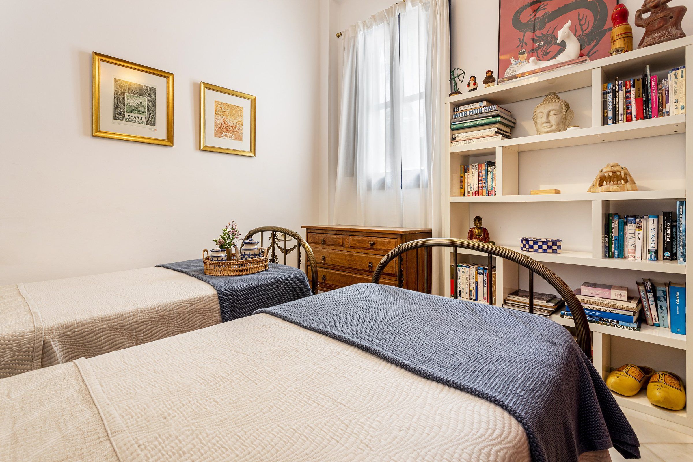 Une chambre avec deux lits, une commode en bois, une bibliothèque remplie de livres et une fenêtre avec des rideaux.