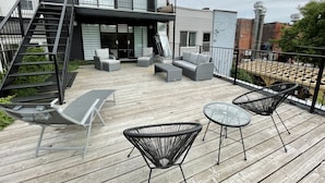 Una terraza de madera con mobiliario exterior, una escalera y un edificio con ventanas amplias y un balcón.