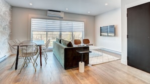 Una sala de estar moderna con un sofá oscuro, una mesa de comedor con sillas de estructura metálica y un ventanal con persianas.