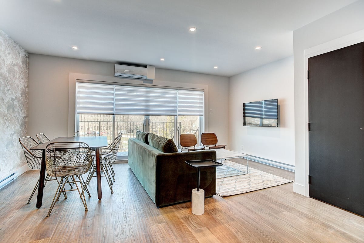 A modern living room with a dark sofa, a dining table with wireframe chairs, and a large window with blinds.