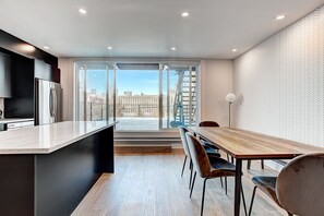 Una cocina moderna con una isla grande, un comedor con una mesa y sillas de madera, y vistas de la ciudad a través de amplias ventanas.