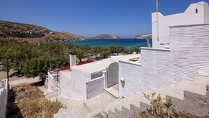 Um edifício costeiro com fachada branca, uma escadaria visível que leva à entrada e vista para o mar e colinas ao longe.