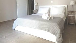 Un lit bien fait, recouvert d’une couverture à carreaux blancs et gris, deux tables de chevet avec des lampes, et une porte donnant sur une autre pièce.