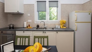 Une cuisine équipée d’une cuisinière, d’un évier et d’un réfrigérateur. Il y a une fenêtre avec des stores, une chaise et une table sur laquelle est posée une citrouille.