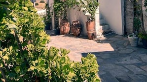 Um pátio estreito com plantas em vasos, um edifício branco com porta e janela, e uma parede de pedra.