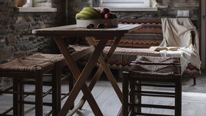 A rustic stone interior with a wooden table, woven chairs, a bench, and a window with a view of the countryside. - Delphis the Artist s House (Tinos)