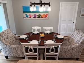 A dining area with a round table, white chairs, and a chandelier. There is a painting of beach chairs on the wall.