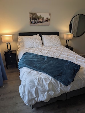 A neatly made bed with white bedding and a blue throw blanket, flanked by two bedside tables with lamps, a framed picture on the wall, and a round mirror. - Paradise Village home in perfect golfing and recreation center location. (The Villages)