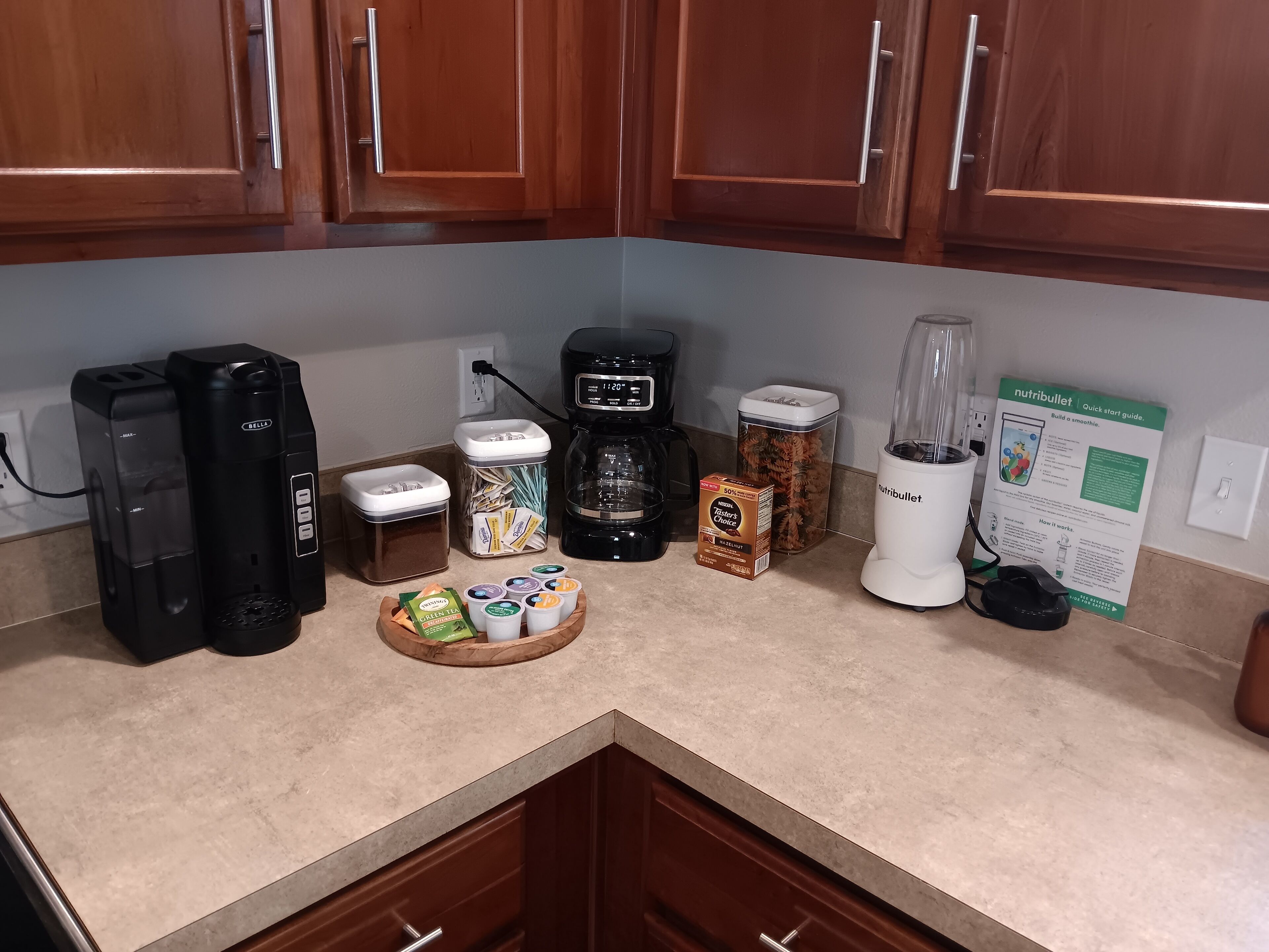 Un plan de travail de cuisine avec une machine à café, un mixeur, un guide d’utilisation du Nutribullet, un bocal de grains de café, une boîte de capsules de café et un bol de sachets de thé.