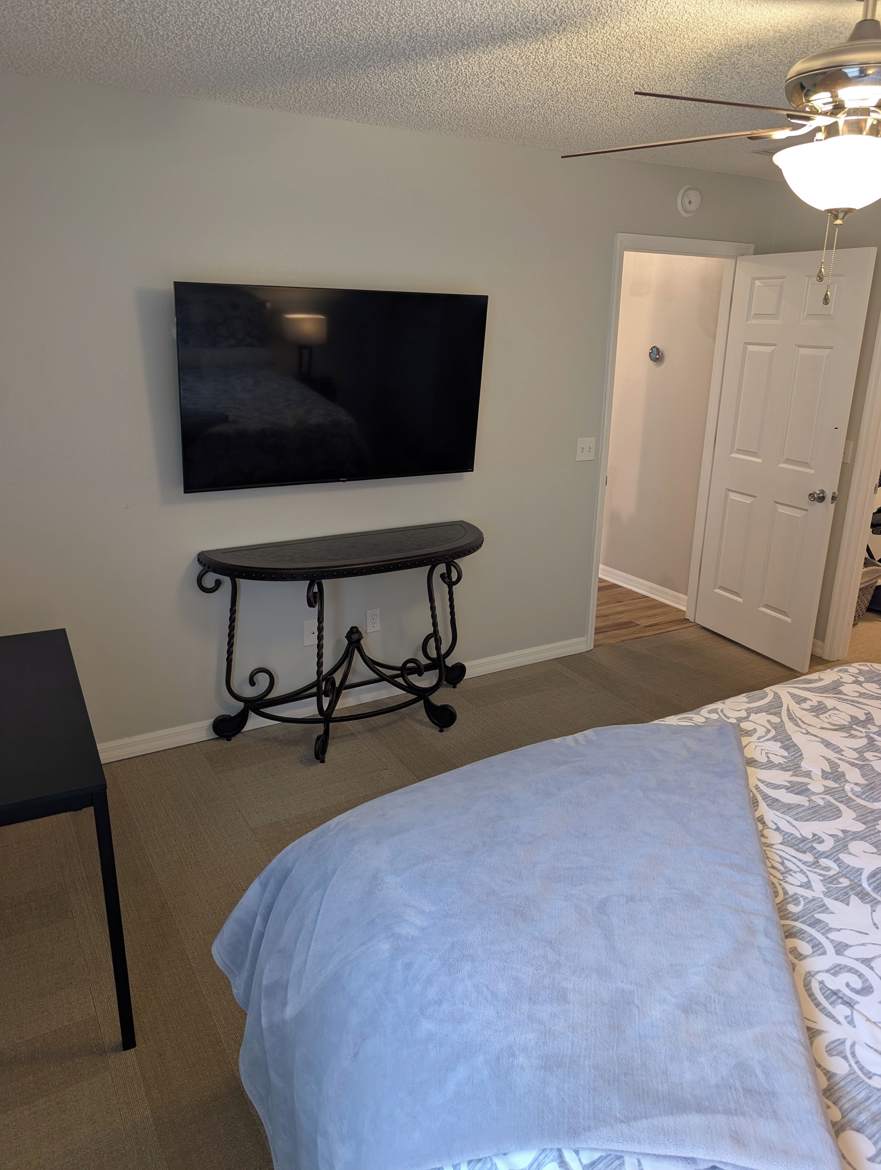 A bedroom with a bed, a TV mounted on the wall, a small table, and a door.