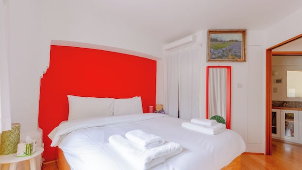 A bedroom with a large bed, white bedding, a red accent wall, a bedside table with a lamp, and a painting on the wall. - Uniquely Designed 2BD Flat near Tower Bridge (London)