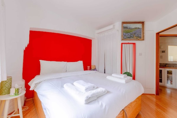 A hotel room with a red accent wall, a bed with white linens, a small table with a teddy bear, and a painting on the wall.