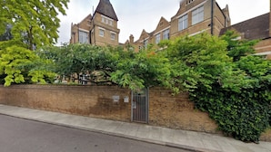 A brick wall with a metal gate, a sidewalk, and a building with multiple windows and a chimney in the background. - Uniquely Designed 2BD Flat near Tower Bridge (London)
