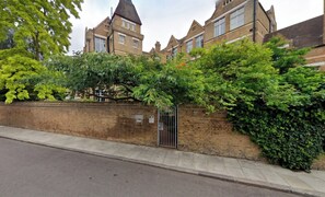 A brick wall with a metal gate, a sidewalk, and a building with multiple windows and a chimney in the background.