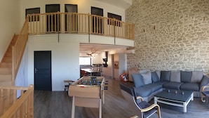 Ein großzügiger Wohnbereich mit einem Kicker, einer Couch und einem Esstisch. Eine Treppe führt zu einer oberen Ebene mit einem Balkon.