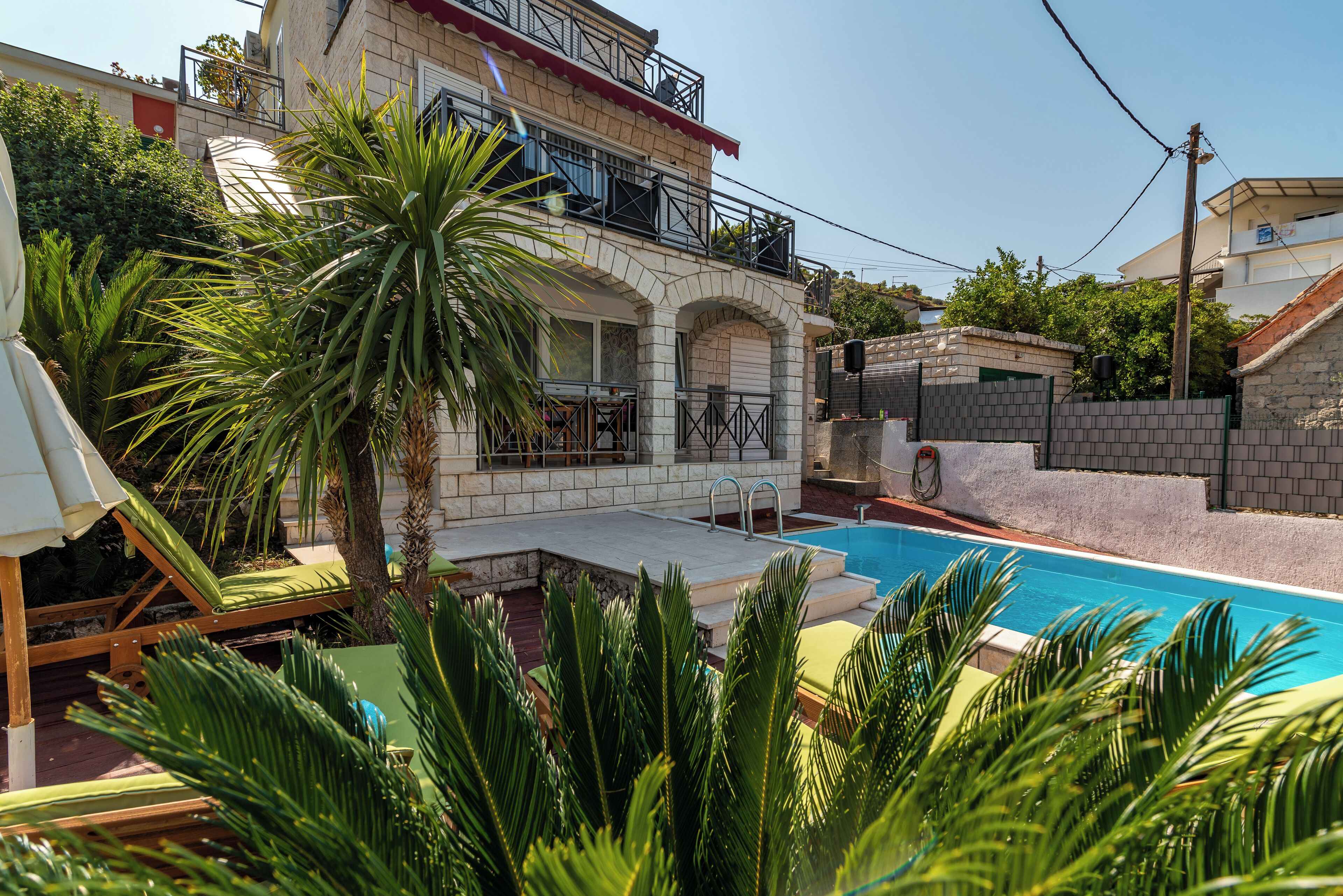 A two-story house with a swimming pool, a balcony, and a garden with palm trees.