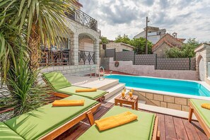 Een zwembadgedeelte met groene loungestoelen en een tafeltje met drankjes, omringd door stenen muren en een palmpijlboom.