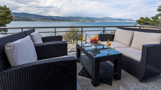 A terrace with wicker furniture and a glass table, overlooking a lake with mountains in the background.