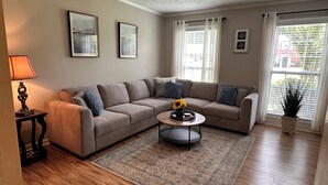 A living room with a beige sofa, a coffee table, and a lamp. There are two framed pictures on the wall and a window with curtains.