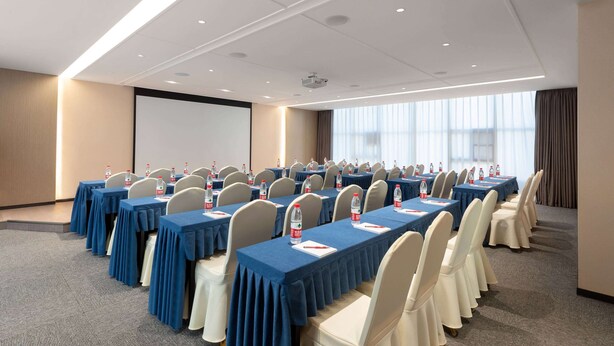 Un salón de conferencias con filas de mesas, sillas y una pantalla de proyector.
