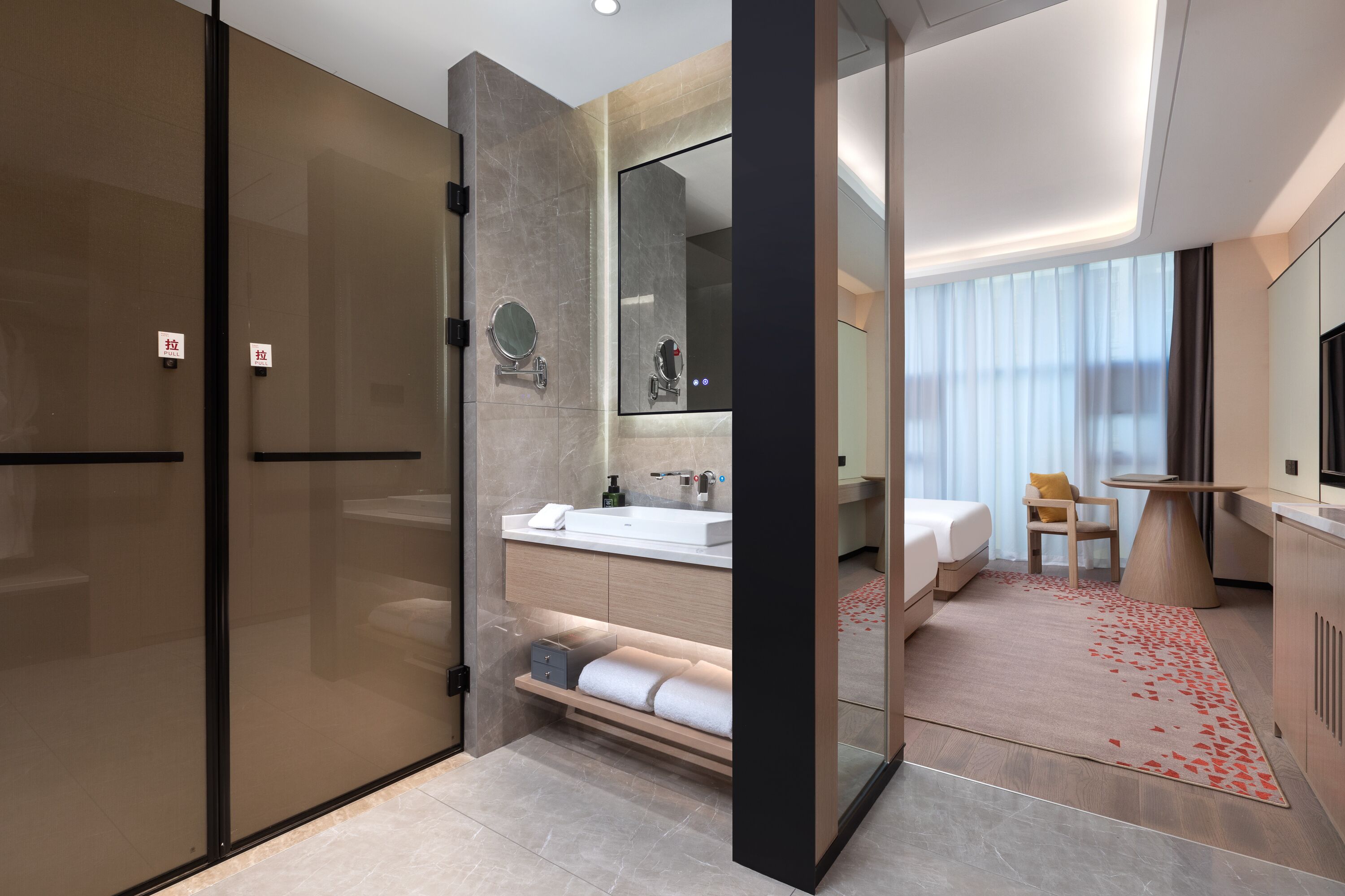 Une chambre d’hôtel moderne dotée d’une douche en verre, d’un grand miroir, d’un lavabo blanc, d’un lit, d’une petite table et d’un tapis rouge orné de motifs floraux.
