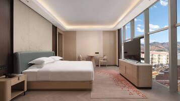 Una habitación de hotel moderna con una cama grande, un escritorio, un televisor y vistas a la ciudad a través de amplias ventanas.