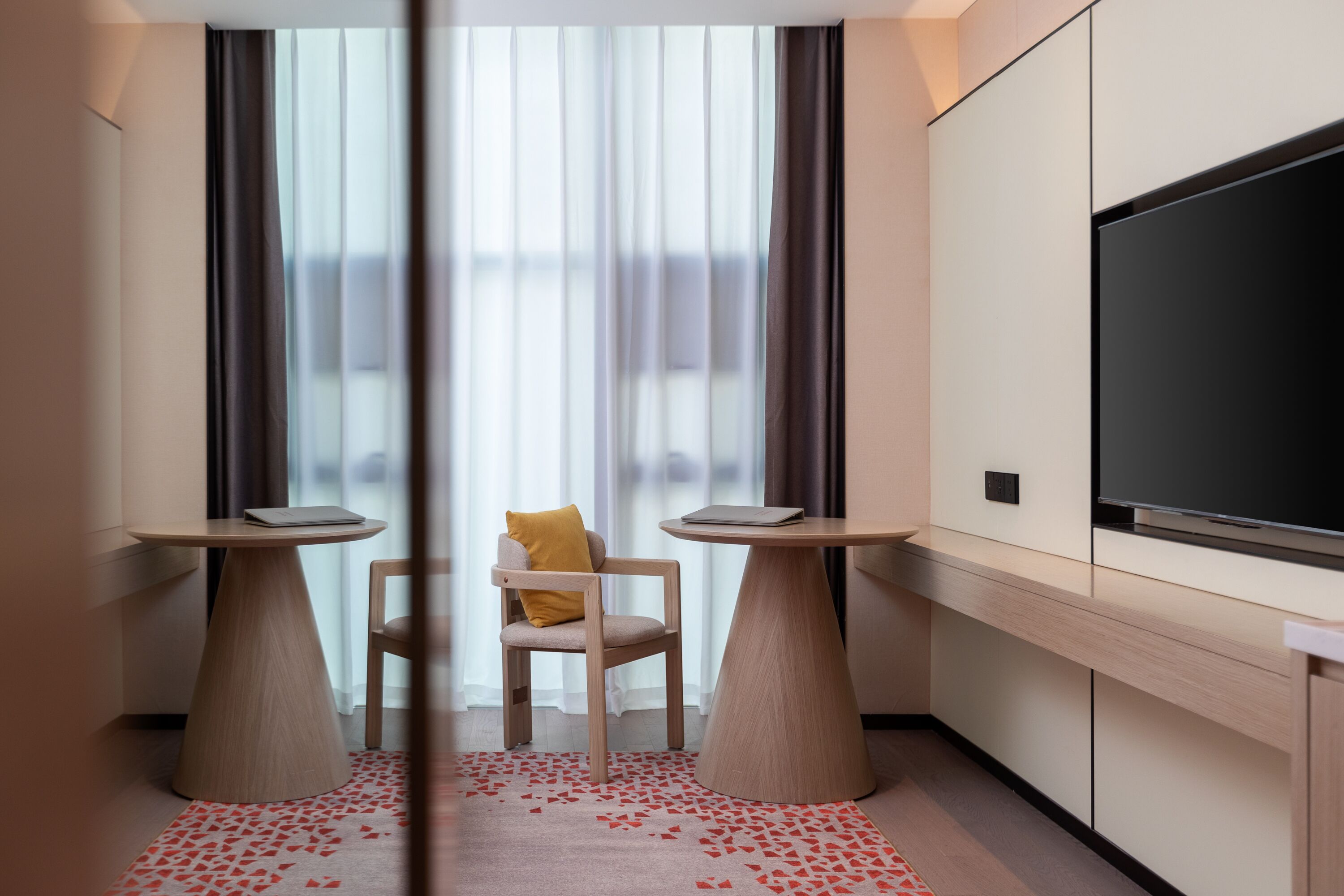 Una habitación de hotel moderna con un televisor de pantalla plana grande, dos mesitas redondas pequeñas, una silla cómoda y un ventanal con cortinas translúcidas.