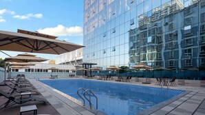 A modern hotel pool area with sun loungers and umbrellas, a clear blue pool, and a glass building reflecting the surroundings. - Ramada by Wyndham Honghe Kaiyuan (Honghe)