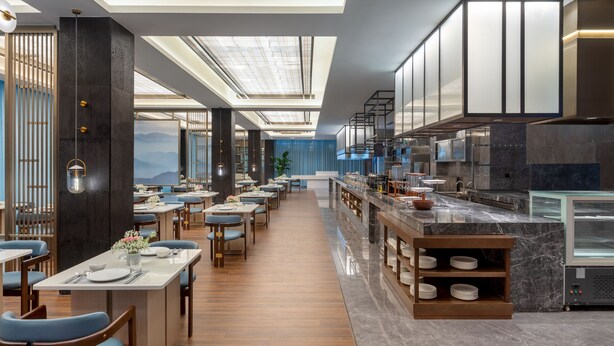 A modern restaurant with a long dining area, a kitchen with marble countertops, and a view of the mountains. - Ramada by Wyndham Honghe Kaiyuan (Honghe)