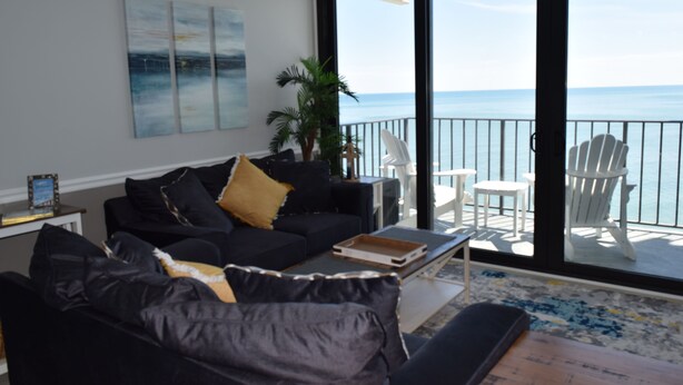 A living room with a black sofa, a coffee table, and a view of the ocean through a sliding glass door. - Oceanfront Family Friendly Garden City Beach Condo (Murrells Inlet)