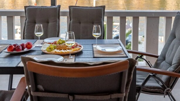 Une table à manger avec des assiettes, des verres et un plateau de fruits, placée sur un balcon donnant sur un lac, avec des maisons en arrière-plan.